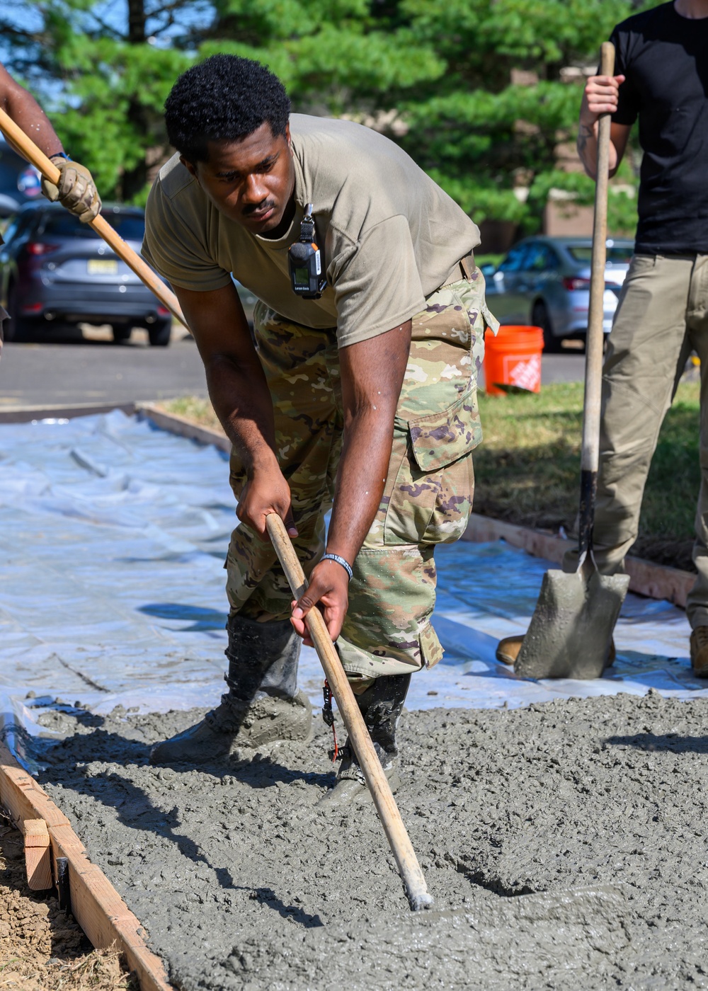 A person wearing military camouflage (OCP/MultiCam) uniform and a tan t-shirt is actively spreading or leveling wet concrete within a wooden form frame. At least two other individuals are visible assisting with the concrete work. The setting appears to be an outdoor construction area near a parking lot with trees in the background.