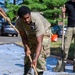 A person wearing military camouflage (OCP/MultiCam) uniform and a tan t-shirt is actively spreading or leveling wet concrete within a wooden form frame. At least two other individuals are visible assisting with the concrete work. The setting appears to be an outdoor construction area near a parking lot with trees in the background.