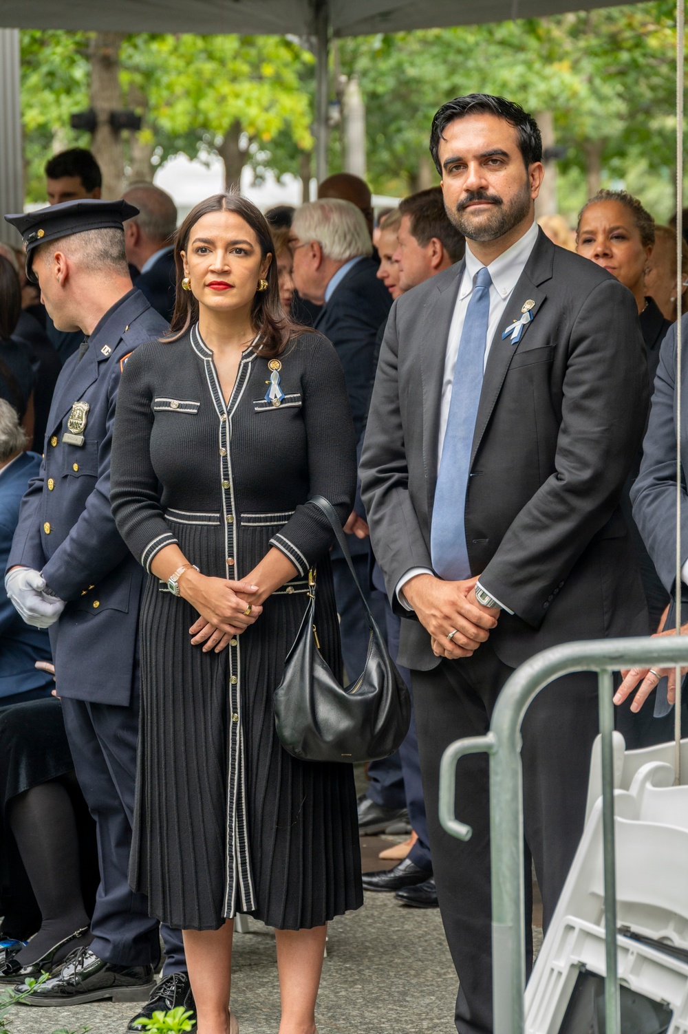 Two formally dressed individuals stand together at what appears to be a solemn outdoor ceremony or memorial event. Both are wearing black attire with blue-and-white ribbon pins on their lapels, suggesting a commemorative occasion. They are surrounded by other attendees including a uniformed law enforcement officer, under a tent with green trees visible in the background.
