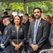 Two formally dressed individuals stand together at what appears to be a solemn outdoor ceremony or memorial event. Both are wearing black attire with blue-and-white ribbon pins on their lapels, suggesting a commemorative occasion. They are surrounded by other attendees including a uniformed law enforcement officer, under a tent with green trees visible in the background.