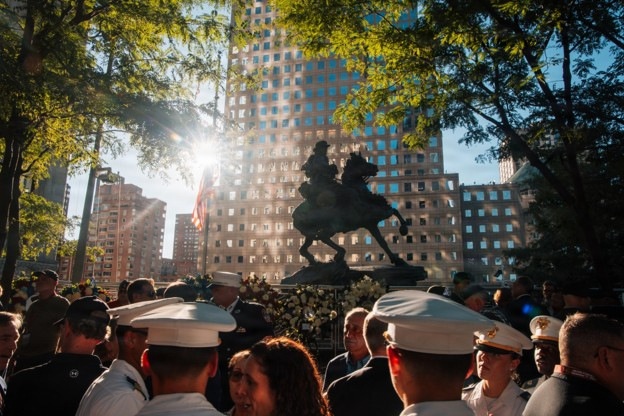 A ceremonial gathering of military and naval personnel surrounds an equestrian statue in an urban plaza, with tall buildings visible in the background. An American flag is visible near the statue, and floral wreaths are placed at the base, suggesting a commemorative or memorial event. The scene is backlit by strong sunlight filtering through trees, creating a dramatic silhouette effect on the statue.