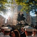 A ceremonial gathering of military and naval personnel surrounds an equestrian statue in an urban plaza, with tall buildings visible in the background. An American flag is visible near the statue, and floral wreaths are placed at the base, suggesting a commemorative or memorial event. The scene is backlit by strong sunlight filtering through trees, creating a dramatic silhouette effect on the statue.