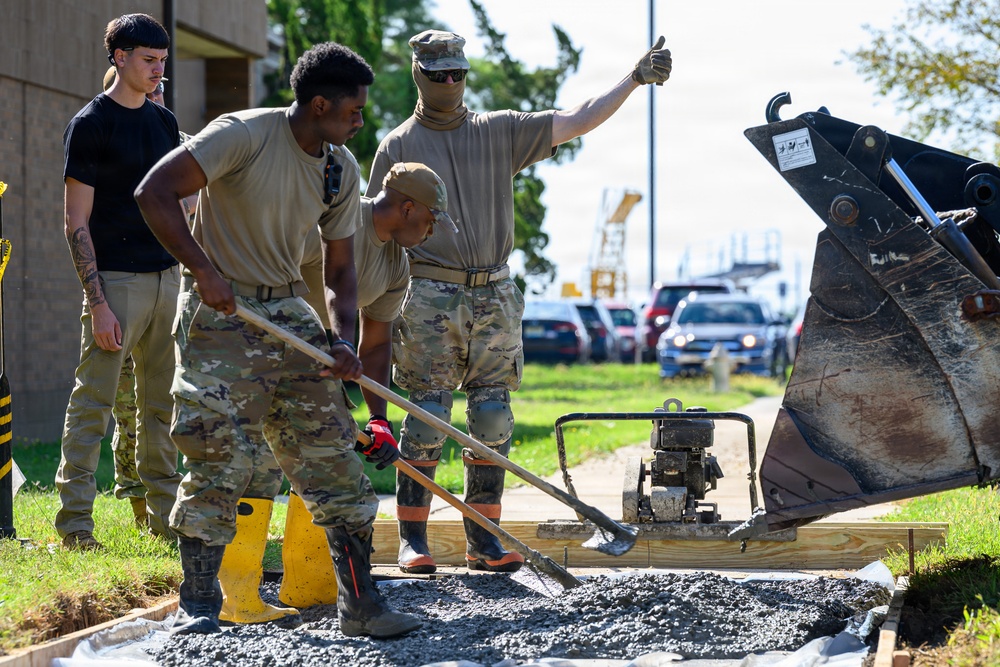 A group of military personnel in camouflage uniforms are performing construction or repair work on a paved surface, spreading gravel or aggregate material with shovels. One individual in a face covering is giving a thumbs-up gesture, while others actively work with tools. A civilian in a black shirt observes nearby, and heavy construction equipment including an excavator bucket is visible on the right.