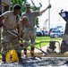 A group of military personnel in camouflage uniforms are performing construction or repair work on a paved surface, spreading gravel or aggregate material with shovels. One individual in a face covering is giving a thumbs-up gesture, while others actively work with tools. A civilian in a black shirt observes nearby, and heavy construction equipment including an excavator bucket is visible on the right.