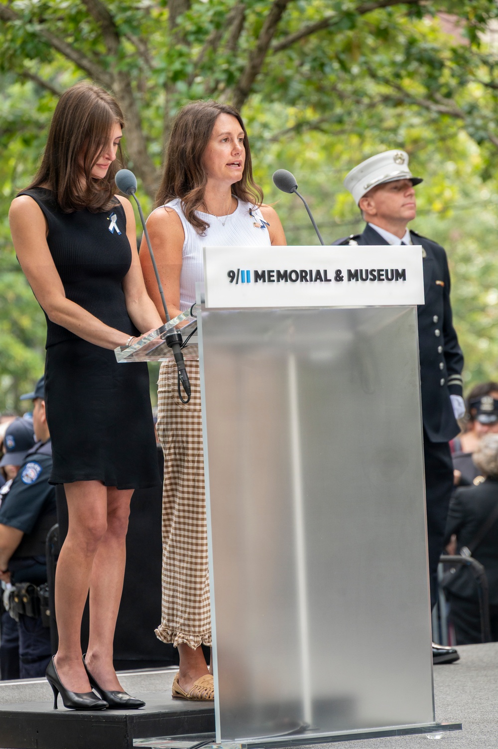 Two women stand at a podium bearing the '9/11 Memorial &amp; Museum' logo during what appears to be a commemorative ceremony. One woman is speaking at the microphone while the other stands beside her holding papers. A uniformed fire department official in a white cap stands in the background.
