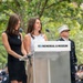 Two women stand at a podium bearing the '9/11 Memorial &amp; Museum' logo during what appears to be a commemorative ceremony. One woman is speaking at the microphone while the other stands beside her holding papers. A uniformed fire department official in a white cap stands in the background.
