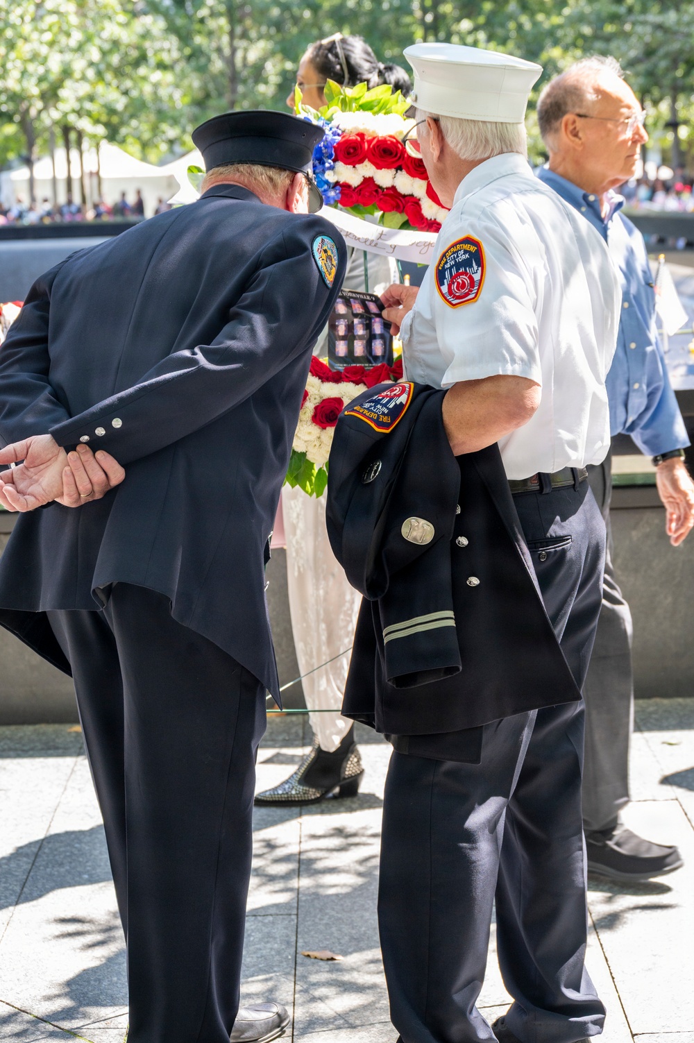 Two FDNY (Fire Department of New York) members in uniform are standing at what appears to be the 9/11 Memorial in New York City. One is wearing a white dress shirt with an FDNY patch and white captain's hat, while the other is in a dark dress uniform. They are participating in a memorial ceremony, with a patriotic red, white, and blue floral wreath visible in the background.
