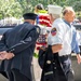 Two FDNY (Fire Department of New York) members in uniform are standing at what appears to be the 9/11 Memorial in New York City. One is wearing a white dress shirt with an FDNY patch and white captain's hat, while the other is in a dark dress uniform. They are participating in a memorial ceremony, with a patriotic red, white, and blue floral wreath visible in the background.