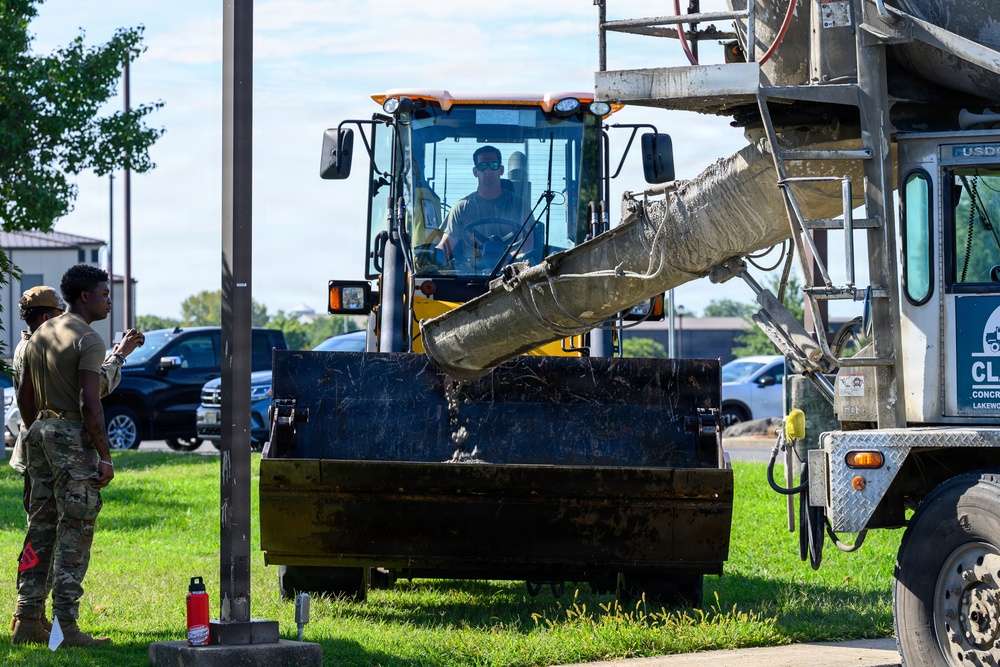 Military personnel in camouflage uniforms are overseeing a construction operation involving a yellow front-end loader and a concrete mixer truck. A civilian operator is seated in the loader cab, positioning the bucket to receive concrete from the mixer chute. The scene appears to take place on a military installation, indicated by the uniforms and facility buildings visible in the background.