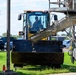 Military personnel in camouflage uniforms are overseeing a construction operation involving a yellow front-end loader and a concrete mixer truck. A civilian operator is seated in the loader cab, positioning the bucket to receive concrete from the mixer chute. The scene appears to take place on a military installation, indicated by the uniforms and facility buildings visible in the background.