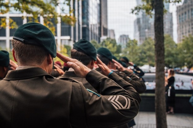A line of military personnel in olive/dark green uniforms and dark green berets are standing at salute near what appears to be a memorial reflecting pool in an urban setting. The soldiers are photographed from behind, facing the memorial. The scene suggests a formal commemorative ceremony, likely at the 9/11 Memorial in New York City based on the distinctive reflecting pool and surrounding skyline.