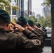 A line of military personnel in olive/dark green uniforms and dark green berets are standing at salute near what appears to be a memorial reflecting pool in an urban setting. The soldiers are photographed from behind, facing the memorial. The scene suggests a formal commemorative ceremony, likely at the 9/11 Memorial in New York City based on the distinctive reflecting pool and surrounding skyline.