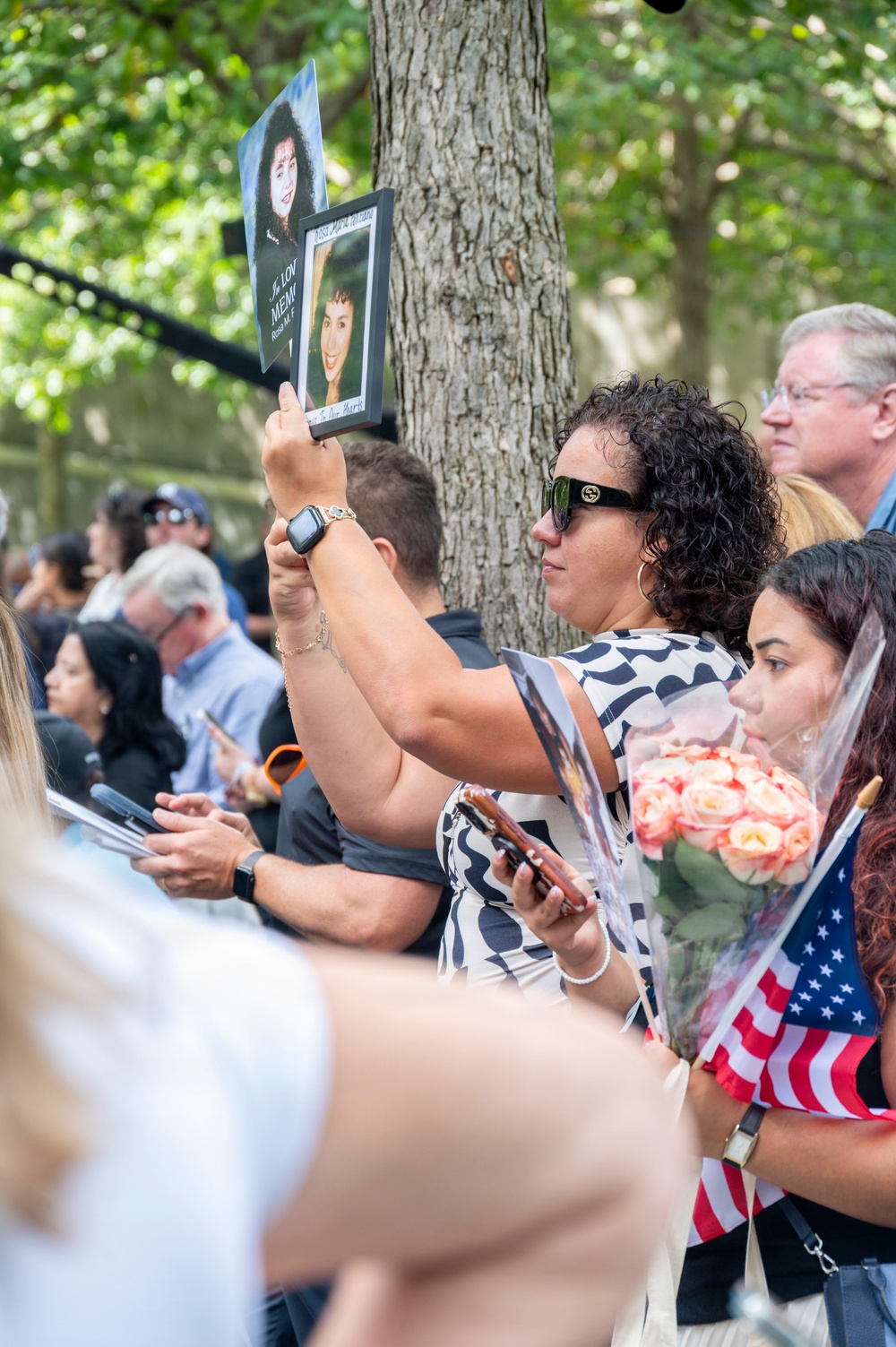 A crowd of people gathered at what appears to be a memorial ceremony, likely a 9/11 anniversary commemoration. A woman in the foreground holds up a framed photograph and a printed photo of a deceased individual, with text partially visible reading 'In Loving Memory' and a name. Other attendees hold flowers, American flags, and personal items in a solemn outdoor setting.
