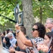 A crowd of people gathered at what appears to be a memorial ceremony, likely a 9/11 anniversary commemoration. A woman in the foreground holds up a framed photograph and a printed photo of a deceased individual, with text partially visible reading 'In Loving Memory' and a name. Other attendees hold flowers, American flags, and personal items in a solemn outdoor setting.