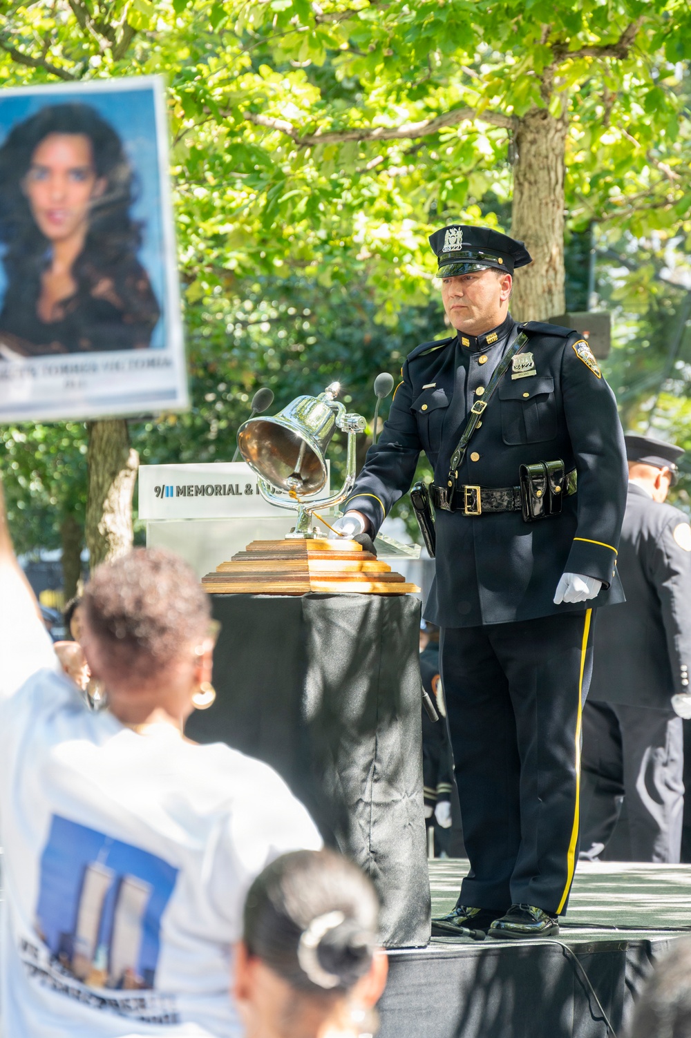 A uniformed NYPD officer in dress blues stands at a podium during a 9/11 Memorial &amp; Museum ceremony, ringing or attending a ceremonial bell mounted on a wooden base. A portrait photograph of a victim is visible on a sign in the background. Attendees, including a person wearing a commemorative Twin Towers t-shirt, are visible in the foreground.