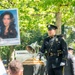 A uniformed NYPD officer in dress blues stands at a podium during a 9/11 Memorial &amp; Museum ceremony, ringing or attending a ceremonial bell mounted on a wooden base. A portrait photograph of a victim is visible on a sign in the background. Attendees, including a person wearing a commemorative Twin Towers t-shirt, are visible in the foreground.