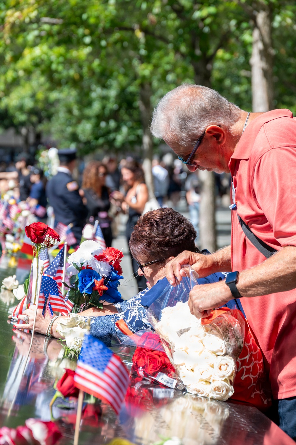 Two older adults are placing flowers and tributes at what appears to be the 9/11 Memorial in New York City. The reflective bronze parapet is lined with American flags, red roses, white roses, and red-white-and-blue floral arrangements. A crowd of visitors and what appears to be a uniformed official are visible in the background among trees.