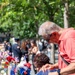 Two older adults are placing flowers and tributes at what appears to be the 9/11 Memorial in New York City. The reflective bronze parapet is lined with American flags, red roses, white roses, and red-white-and-blue floral arrangements. A crowd of visitors and what appears to be a uniformed official are visible in the background among trees.