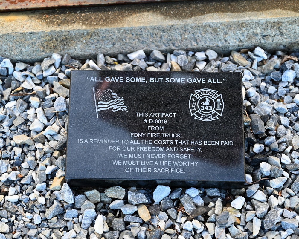 A black granite memorial plaque resting on gravel, commemorating the September 11, 2001 attacks. The plaque identifies an artifact (# D-0016) from an FDNY fire truck and bears the quote 'All gave some, but some gave all.' It features engraved imagery of an American flag and a firefighter memorial emblem referencing the 343 FDNY firefighters lost on 9/11.