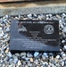 A black granite memorial plaque resting on gravel, commemorating the September 11, 2001 attacks. The plaque identifies an artifact (# D-0016) from an FDNY fire truck and bears the quote 'All gave some, but some gave all.' It features engraved imagery of an American flag and a firefighter memorial emblem referencing the 343 FDNY firefighters lost on 9/11.
