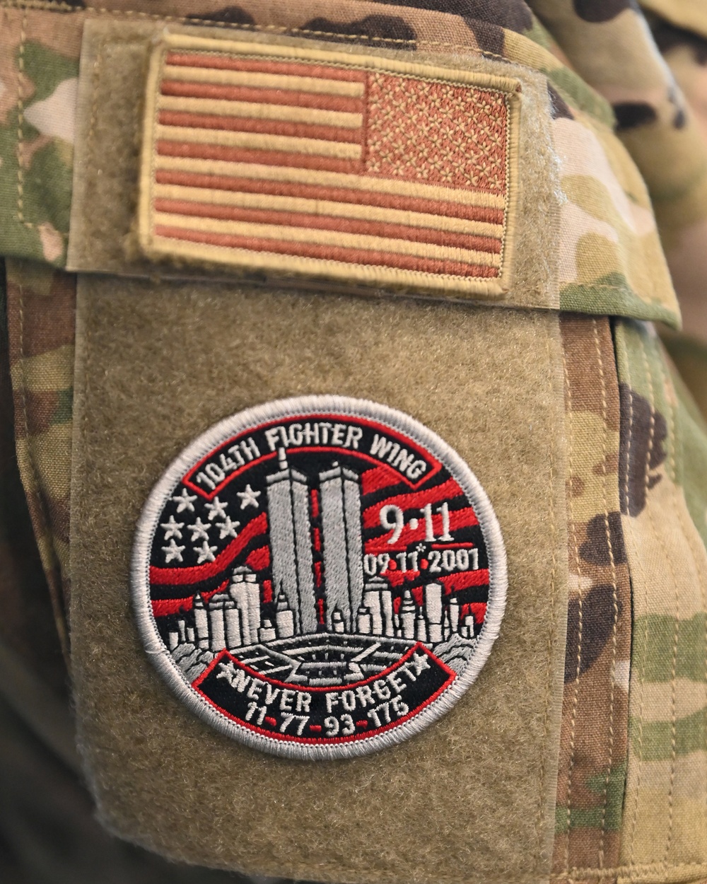 The image shows the upper arm/shoulder area of a military uniform in MultiCam/OCP camouflage pattern. Two patches are visible: a subdued U.S. flag patch on the upper portion and a commemorative 9/11 memorial patch below it. The memorial patch belongs to the 104th Fighter Wing and honors the September 11, 2001 attacks with the motto 'Never Forget' and the numbers 11-77-93-175.