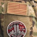 The image shows the upper arm/shoulder area of a military uniform in MultiCam/OCP camouflage pattern. Two patches are visible: a subdued U.S. flag patch on the upper portion and a commemorative 9/11 memorial patch below it. The memorial patch belongs to the 104th Fighter Wing and honors the September 11, 2001 attacks with the motto 'Never Forget' and the numbers 11-77-93-175.