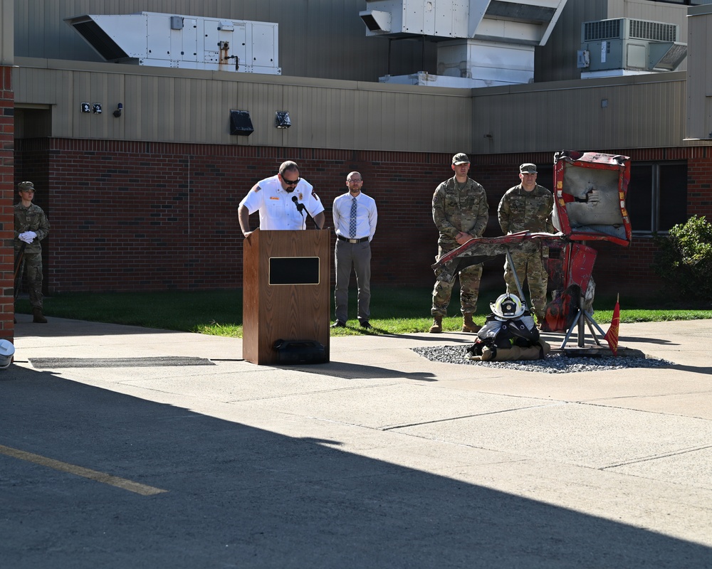 An outdoor ceremony is taking place in front of a brick and metal building, likely on a military installation. A person in a white fire department uniform is speaking at a wooden podium, while military personnel in camouflage uniforms and a civilian stand in attendance. A memorial display featuring burned/damaged fire truck components, firefighter gear, and a small red flag is prominently positioned to the right.