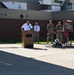 An outdoor ceremony is taking place in front of a brick and metal building, likely on a military installation. A person in a white fire department uniform is speaking at a wooden podium, while military personnel in camouflage uniforms and a civilian stand in attendance. A memorial display featuring burned/damaged fire truck components, firefighter gear, and a small red flag is prominently positioned to the right.