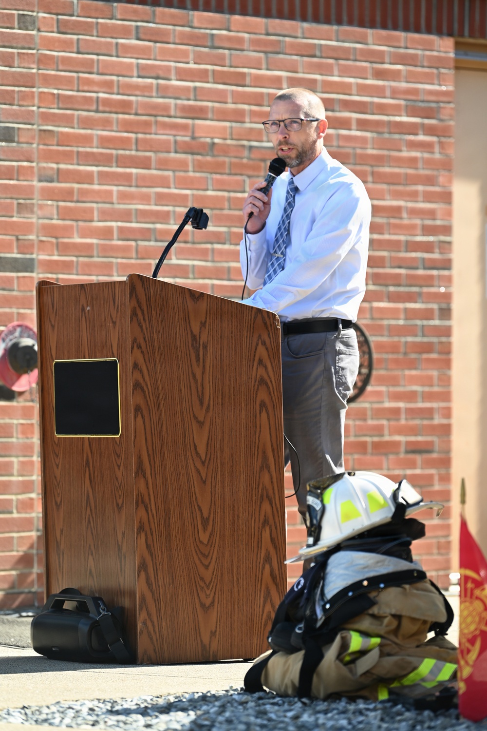 A man in business casual attire (white dress shirt, tie, gray slacks) is speaking at a wooden podium while holding a handheld microphone outdoors. The setting appears to be a ceremonial or commemorative event at a fire station, evidenced by firefighting gear (helmet, turnout coat) placed on the ground in the foreground and a red fire department flag visible to the right.