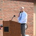 A man in business casual attire (white dress shirt, tie, gray slacks) is speaking at a wooden podium while holding a handheld microphone outdoors. The setting appears to be a ceremonial or commemorative event at a fire station, evidenced by firefighting gear (helmet, turnout coat) placed on the ground in the foreground and a red fire department flag visible to the right.