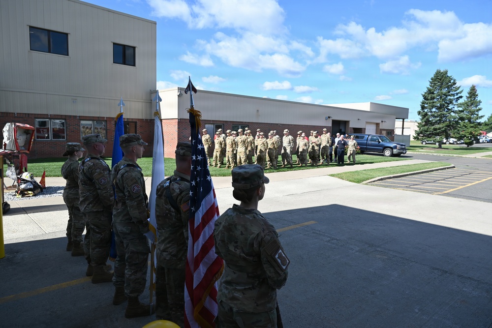 A military color guard ceremony is taking place outdoors at what appears to be a military installation or National Guard facility. Several soldiers in Army Combat Uniforms (ACU/OCP pattern) are holding flags including the United States flag and at least one unit or service flag, while a formation of uniformed personnel stands at attention in the background. The event appears to be a formal military ceremony such as a change of command, activation, or unit recognition event.