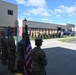 A military color guard ceremony is taking place outdoors at what appears to be a military installation or National Guard facility. Several soldiers in Army Combat Uniforms (ACU/OCP pattern) are holding flags including the United States flag and at least one unit or service flag, while a formation of uniformed personnel stands at attention in the background. The event appears to be a formal military ceremony such as a change of command, activation, or unit recognition event.