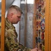 A U.S. military service member in OCP (Operational Camouflage Pattern) uniform is placing or arranging a patch on a wooden display case. The case contains numerous military and fire protection patches mounted on a board behind glass. One clearly visible patch reads 'FIRE PROTECTION USAF' with a circular design.