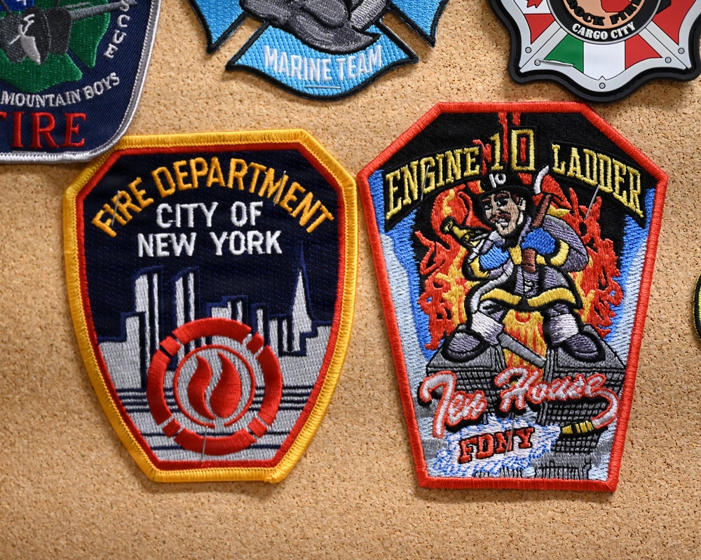 The image shows a collection of embroidered patches pinned to a cork board. The two most prominent patches are from the FDNY (Fire Department City of New York), including a standard FDNY shield patch and an Engine 10 Ladder 10 'Ten House' patch. Additional patches are partially visible around the edges, including a 'Marine Team' patch and others.