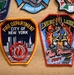 The image shows a collection of embroidered patches pinned to a cork board. The two most prominent patches are from the FDNY (Fire Department City of New York), including a standard FDNY shield patch and an Engine 10 Ladder 10 'Ten House' patch. Additional patches are partially visible around the edges, including a 'Marine Team' patch and others.