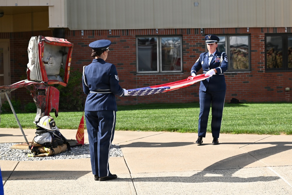Two U.S. Air Force personnel in dress blue uniforms are performing a flag folding ceremony outdoors in front of a brick building. A memorial display is visible to the left, featuring what appears to be twisted metal debris and firefighter gear, suggesting a 9/11 commemorative ceremony. The scene takes place on a paved area with a well-maintained lawn in the background.