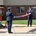 Two U.S. Air Force personnel in dress blue uniforms are performing a flag folding ceremony outdoors in front of a brick building. A memorial display is visible to the left, featuring what appears to be twisted metal debris and firefighter gear, suggesting a 9/11 commemorative ceremony. The scene takes place on a paved area with a well-maintained lawn in the background.