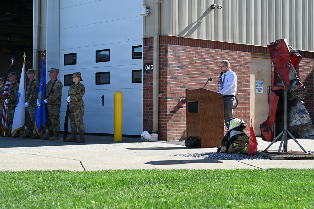 An outdoor ceremony is taking place in front of a military or government building, likely a fire station (Building 040). A civilian speaker addresses the gathering from a wooden podium, while a color guard of uniformed military personnel stands at attention holding flags. A twisted piece of red metal debris — possibly a 9/11 memorial artifact — is displayed on a stand near the podium, alongside firefighter gear.