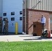 An outdoor ceremony is taking place in front of a military or government building, likely a fire station (Building 040). A civilian speaker addresses the gathering from a wooden podium, while a color guard of uniformed military personnel stands at attention holding flags. A twisted piece of red metal debris — possibly a 9/11 memorial artifact — is displayed on a stand near the podium, alongside firefighter gear.