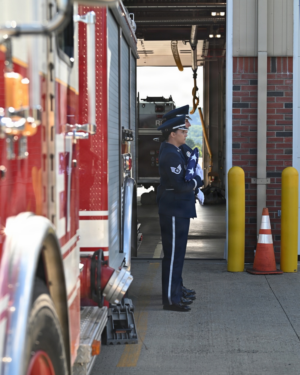Two U.S. Air Force honor guard members in dress blue uniforms stand at attention outside a fire station bay, with one visibly holding a folded American flag. A red fire truck is parked to the left, and the setting appears to be a ceremonial occasion at a military or civilian fire station. The scene suggests a formal tribute or memorial ceremony.