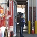 Two U.S. Air Force honor guard members in dress blue uniforms stand at attention outside a fire station bay, with one visibly holding a folded American flag. A red fire truck is parked to the left, and the setting appears to be a ceremonial occasion at a military or civilian fire station. The scene suggests a formal tribute or memorial ceremony.