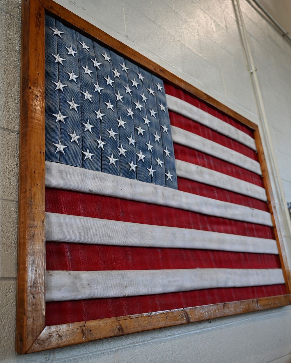 A decorative American flag artwork is mounted on a cinder block wall, framed in a rustic wooden frame. The flag is constructed from textured fabric or rubber-like material with raised three-dimensional stars on the blue canton and alternating red and white stripes. The piece appears to be a handcrafted or artisan-made wall display.