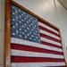 A decorative American flag artwork is mounted on a cinder block wall, framed in a rustic wooden frame. The flag is constructed from textured fabric or rubber-like material with raised three-dimensional stars on the blue canton and alternating red and white stripes. The piece appears to be a handcrafted or artisan-made wall display.