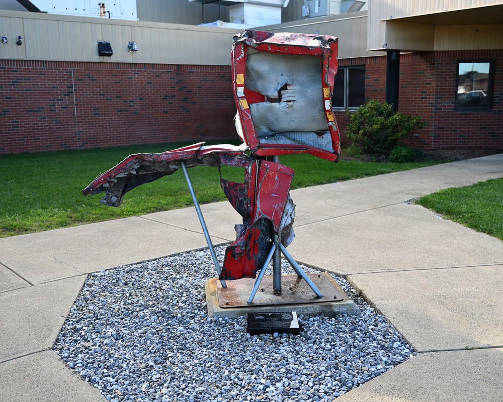 An outdoor metal sculpture made from crushed and distorted red vehicle parts is displayed on a tripod-style metal stand mounted on a base plate. The sculpture is positioned on a gravel bed within a concrete octagonal surround, situated in front of a brick building. A small commemorative plaque is visible at the base of the sculpture.