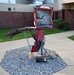 An outdoor metal sculpture made from crushed and distorted red vehicle parts is displayed on a tripod-style metal stand mounted on a base plate. The sculpture is positioned on a gravel bed within a concrete octagonal surround, situated in front of a brick building. A small commemorative plaque is visible at the base of the sculpture.