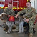 Two U.S. Air Force personnel in OCP (Operational Camouflage Pattern) uniforms pose with a First Responder Therapy Dog inside what appears to be a fire station. The dog, a wire-haired pointing griffon or similar breed, is wearing a red vest labeled 'First Responder Therapy Dog.' A red fire truck is visible in the background with a U.S. Government license plate reading 'AF14C00084'.