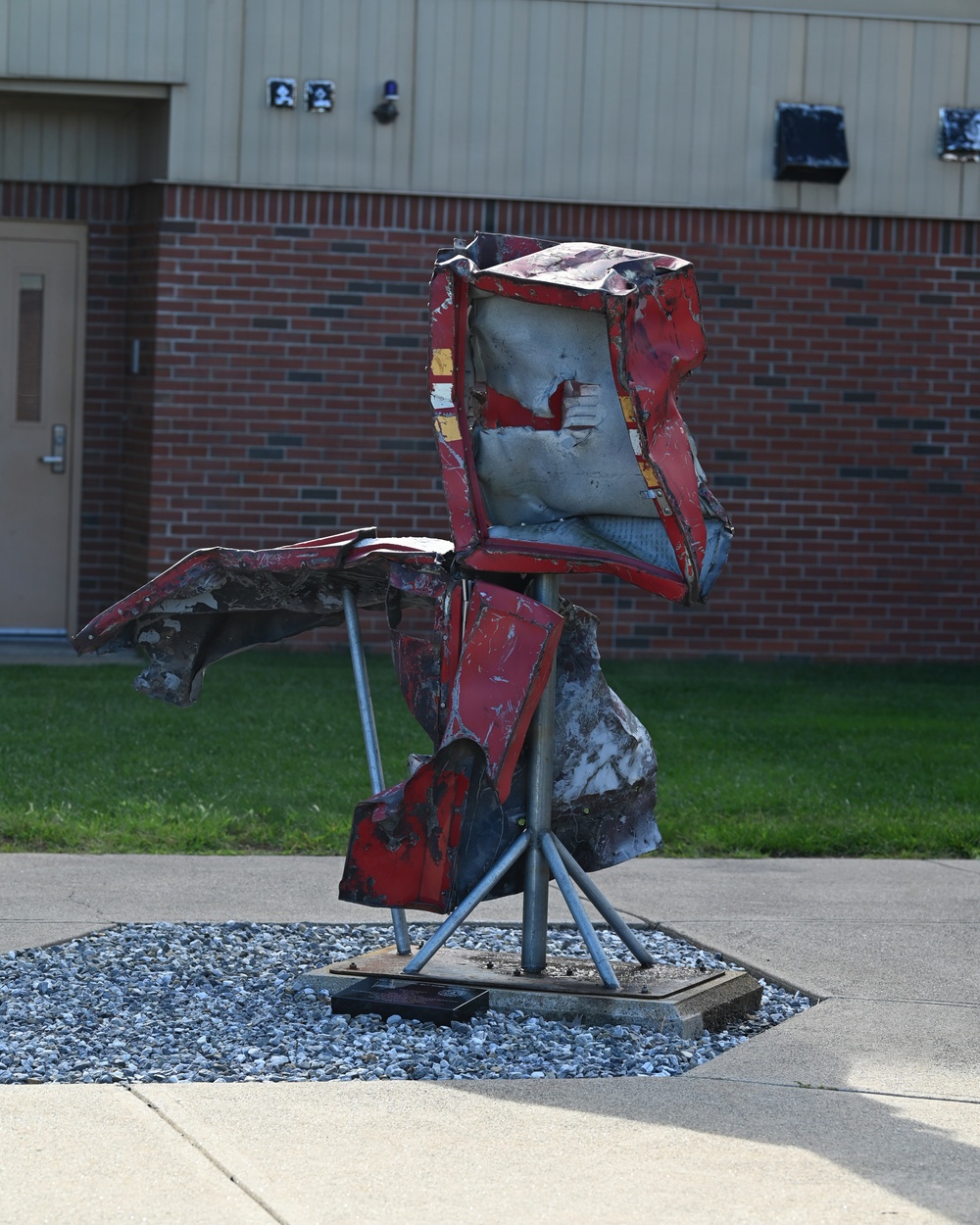 An outdoor metal sculpture composed of crushed and twisted red automotive body panels mounted on galvanized steel pipe supports, displayed on a hexagonal gravel-filled base in front of a brick building. The sculpture appears to be made from salvaged vehicle wreckage, arranged in an abstract humanoid or bird-like form. A small placard or dedication plate is visible at the base of the sculpture.