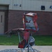 An outdoor metal sculpture composed of crushed and twisted red automotive body panels mounted on galvanized steel pipe supports, displayed on a hexagonal gravel-filled base in front of a brick building. The sculpture appears to be made from salvaged vehicle wreckage, arranged in an abstract humanoid or bird-like form. A small placard or dedication plate is visible at the base of the sculpture.