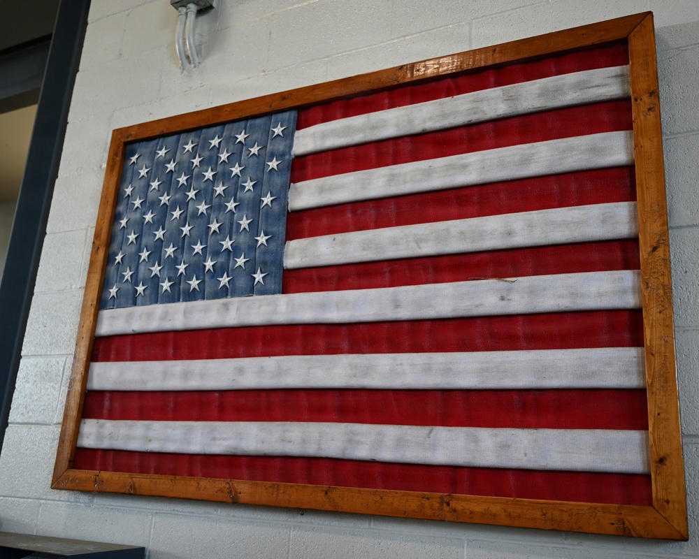 A decorative American flag artwork is mounted on a white cinder block wall, framed in a rustic wooden frame. The flag appears to be constructed from corrugated metal or painted wood planks with raised three-dimensional stars on the blue canton. The piece has a distressed, weathered aesthetic typical of rustic or farmhouse-style décor.