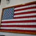 A decorative American flag artwork is mounted on a white cinder block wall, framed in a rustic wooden frame. The flag appears to be constructed from corrugated metal or painted wood planks with raised three-dimensional stars on the blue canton. The piece has a distressed, weathered aesthetic typical of rustic or farmhouse-style décor.